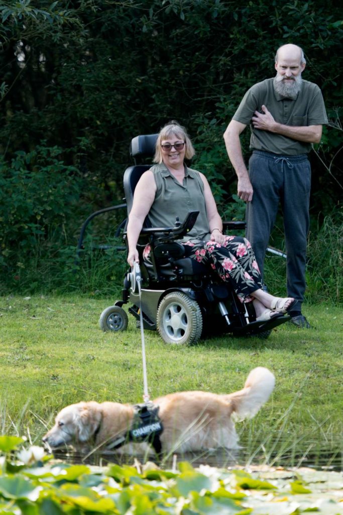 Kvinde i kørestol med hund i snor og en mand gående ved siden af.